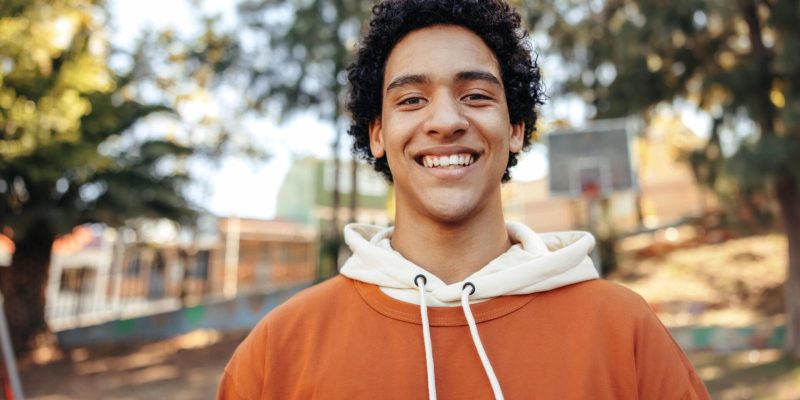 Happy male youngster smiling at the camera outdoors. Fashionable teenager wearing casual clothing in an urban park. Cheerful teenage boy standing alone during the day.